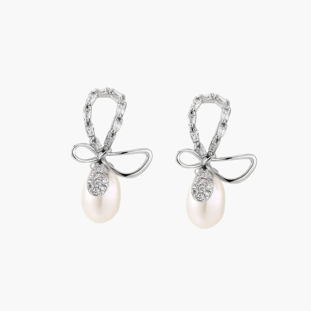 Silver bow-shaped stud earrings with baguette crystals and dangling white freshwater pearls on cream background